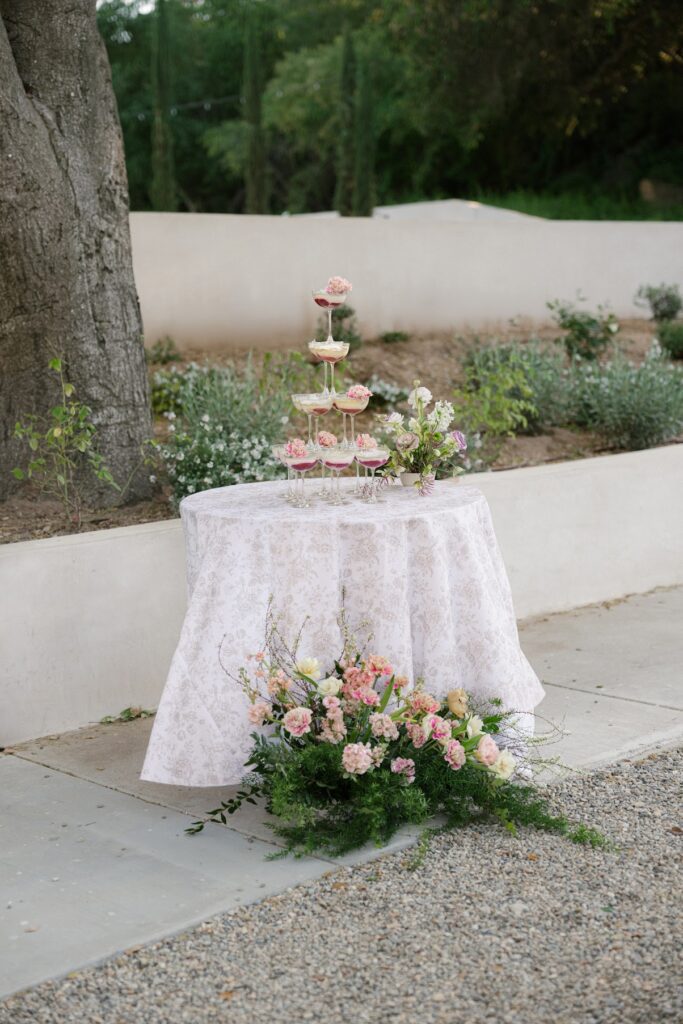 A tiered display of coupe glasses filled with colorful match tiramisu and pink floral garnishes sits on a patterned white tablecloth, surrounded by pastel floral arrangements in an elegant outdoor garden setting at Villa Vienna.