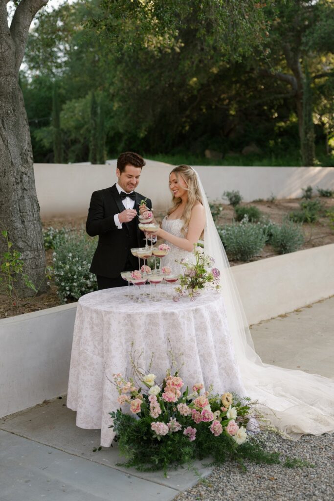 A bride in a strapless lace gown and veil stands beside her groom in a black tuxedo as they pose next to a round table featuring a tiered tower of coupe glasses garnished with pink flowers, set in a lush outdoor garden setting at Villa Vienna.