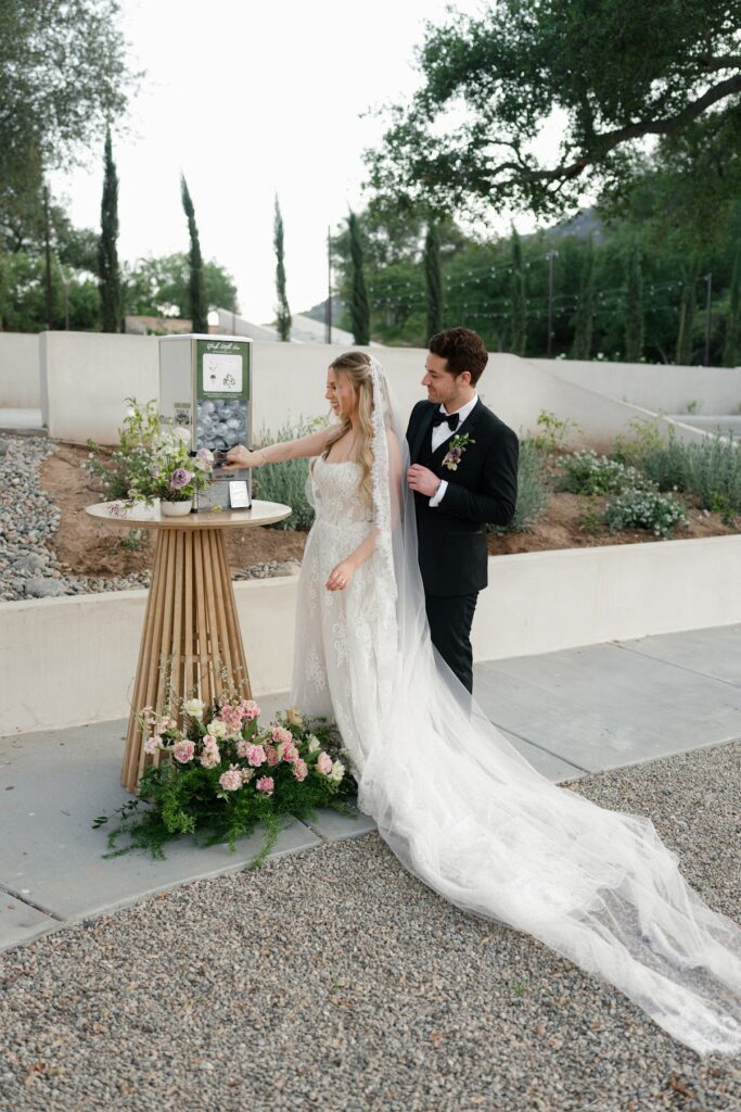 A bride in a lace gown and veil turns the knob of a capsule vending machine filled with sticker filled capsules while her groom in a black tuxedo stands beside her, surrounded by floral arrangements in an outdoor garden setting.