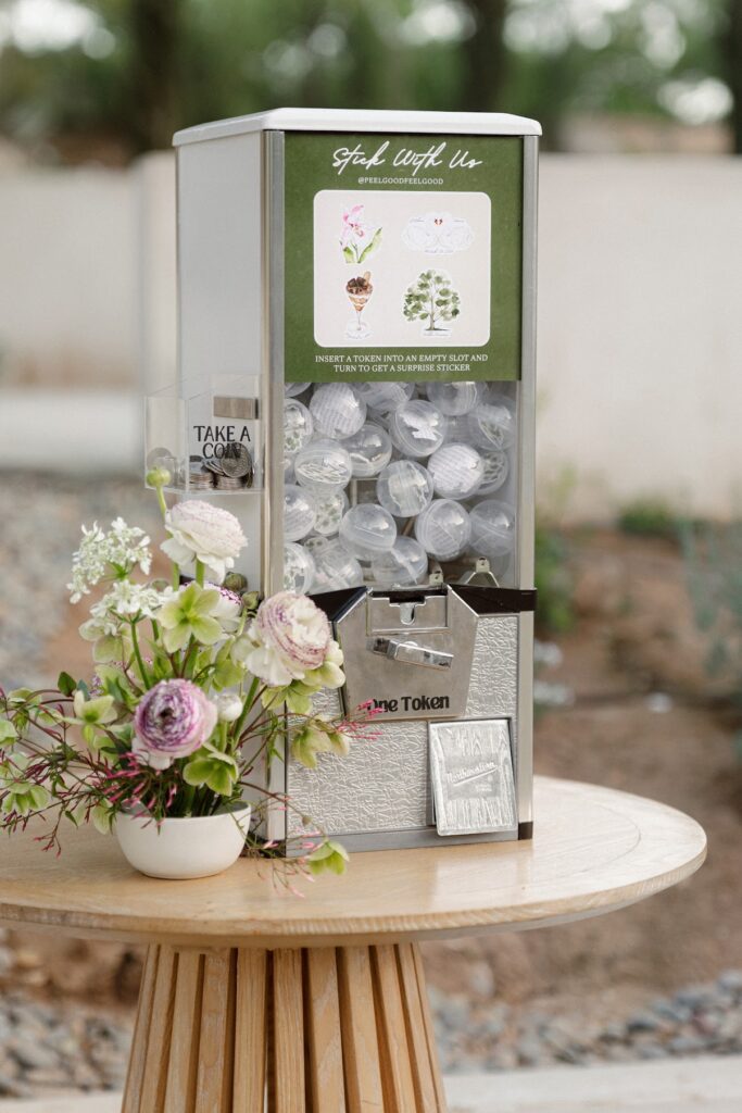 A capsule‑style sticker vending machine sits on a round wooden table outdoors, filled with clear capsules and displayed alongside a small pastel floral arrangement, creating a playful and personalized wedding guest experience.