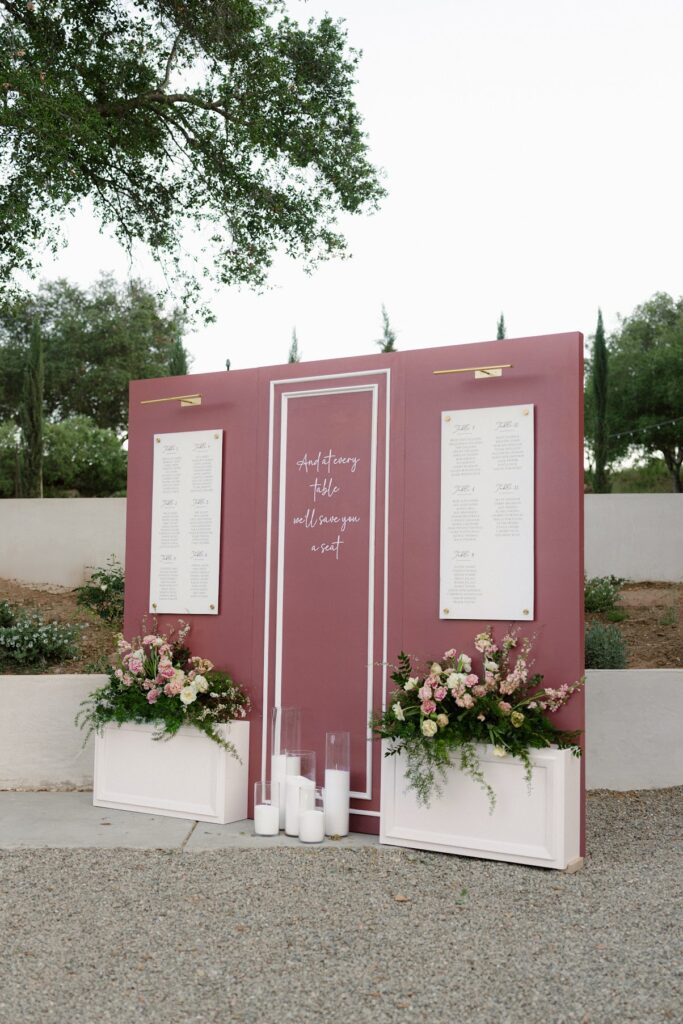 A mauve wedding seating chart display with white table‑assignment panels, gold hardware, and lush pink and cream floral arrangements at the base, accented by tall glass candles, set outdoors among greenery and trees.