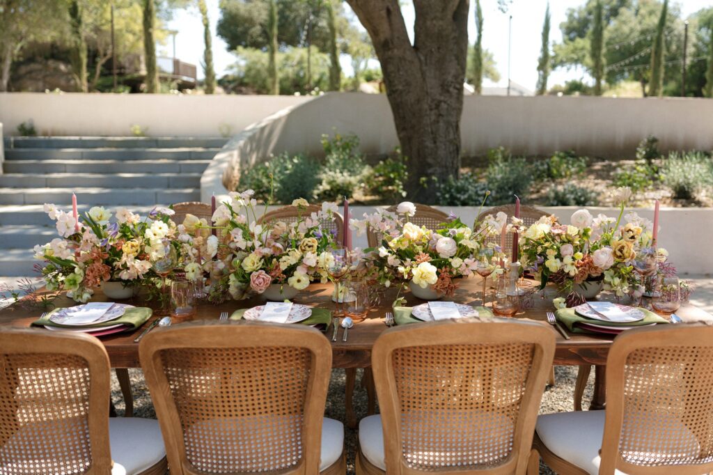 A romantic outdoor wedding tablescape featuring a wooden table set with pastel floral centerpieces, pink taper candles, floral‑patterned plates, green napkins, and elegant glassware, styled in a European‑inspired garden setting at Villa Vienna.