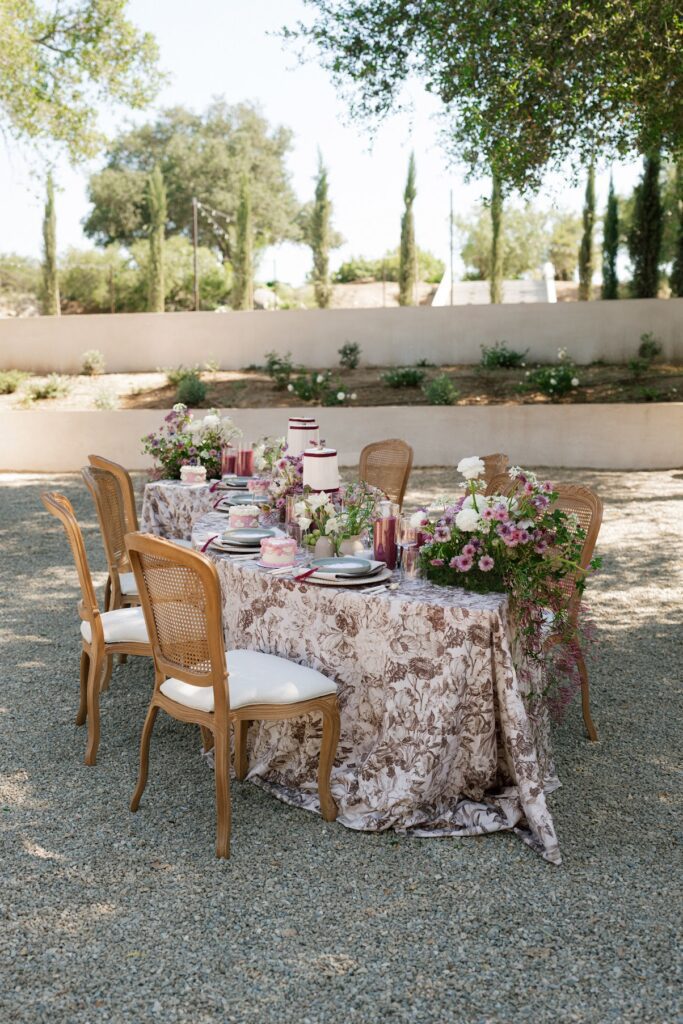 An outdoor serpentine wedding table covered in a neutral floral linen, set with elegant place settings, pastel floral centerpieces, and small decorative cakes, surrounded by wooden chairs in a garden courtyard with cypress trees in the distance at Villa Vienna.