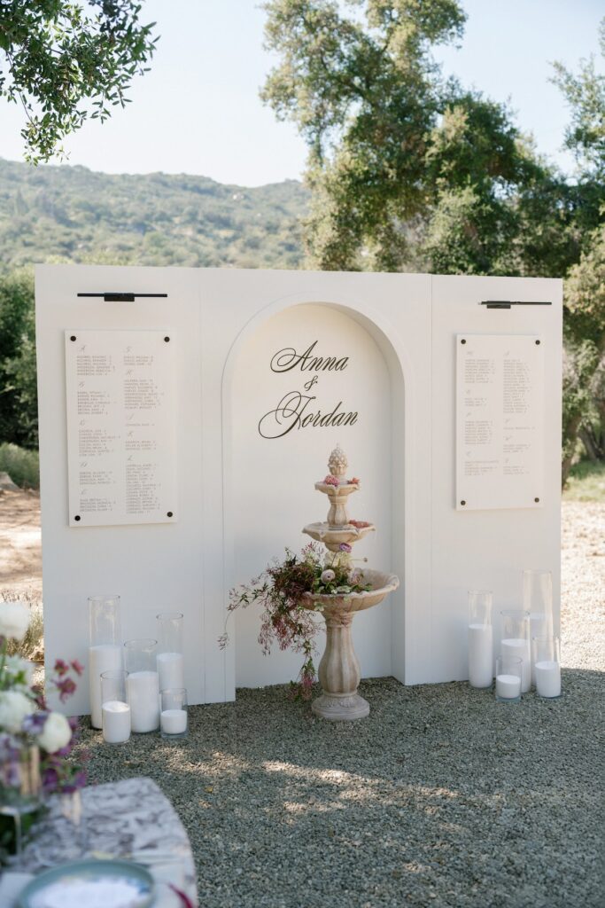 A white wedding seating chart display featuring an arched center panel with the couple’s names above a floral‑accented stone fountain, flanked by two white alphabetical seating panels, surrounded by candles and set outdoors among trees and hills.