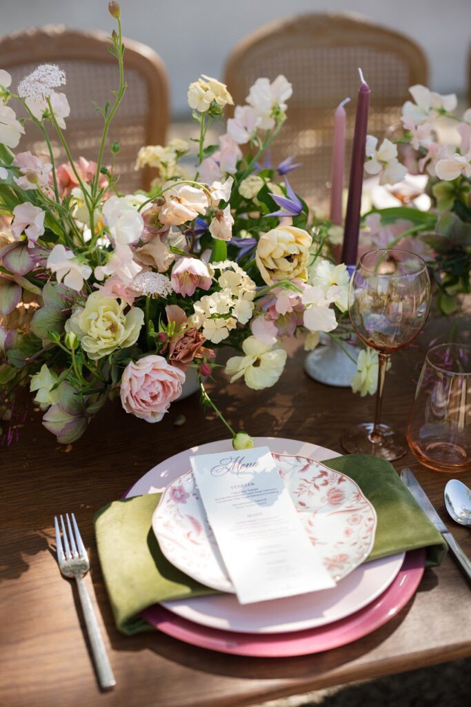 A romantic outdoor wedding tablescape featuring a wooden table set with pastel floral centerpieces, pink taper candles, floral‑patterned plates, green napkins, and elegant glassware, styled in a European‑inspired garden setting at Villa Vienna.