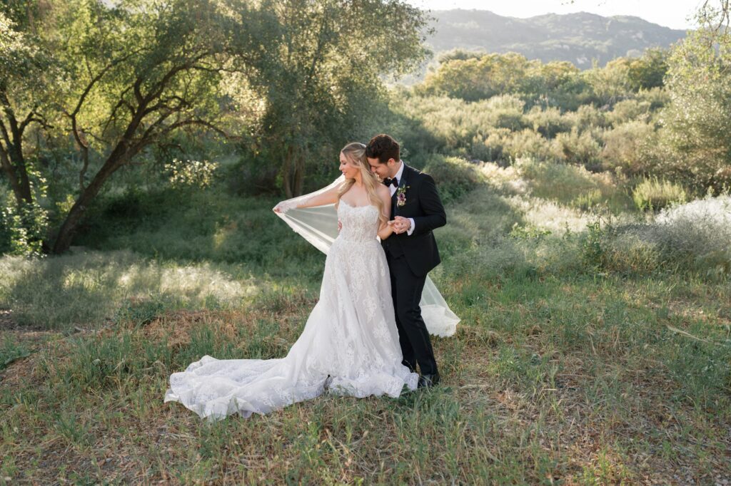 A bride in a lace gown and veil stands hand‑in‑hand with her groom in a black suit, posing together in a lush outdoor setting surrounded by tall trees, greenery, and soft sunlight.