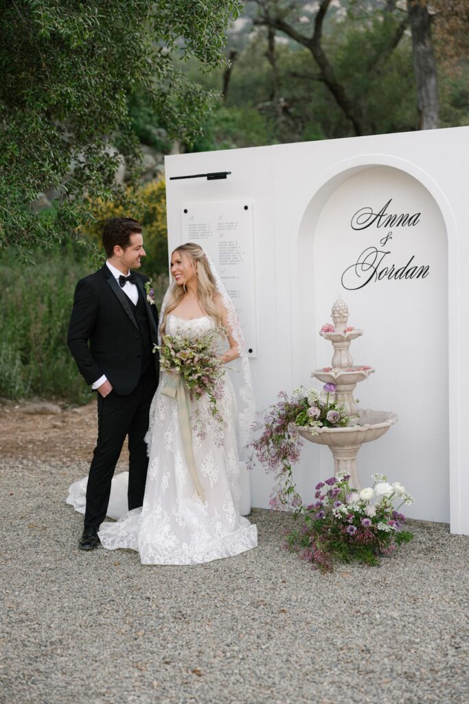 A bride in a lace gown and veil stands beside her groom in a black tuxedo in front of a white arched seating chart wall with “Anna & Jordan” displayed above a floral‑decorated stone fountain, set in a natural outdoor garden setting.