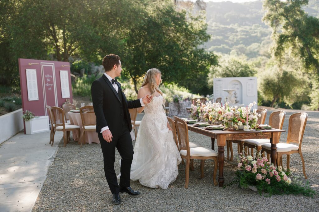 A bride in a lace gown holds hands with her groom beside a long wooden reception table adorned with pastel florals at Villa Vienna, with a mauve seating chart display and fountain installation visible in the background of the outdoor garden setting.