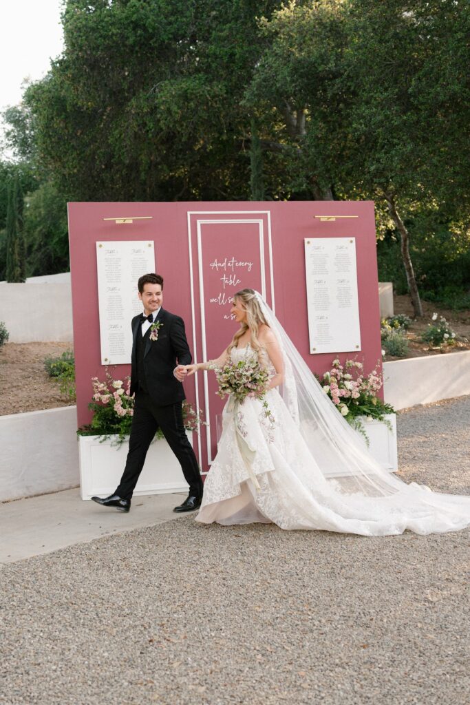 A bride in a lace gown and veil walks hand‑in‑hand with her groom in a black tuxedo past a mauve seating chart backdrop featuring white panels, floral arrangements at the base, and the phrase “And at every table we’ll save a seat,” set in a lush outdoor garden at Villa Vienna.