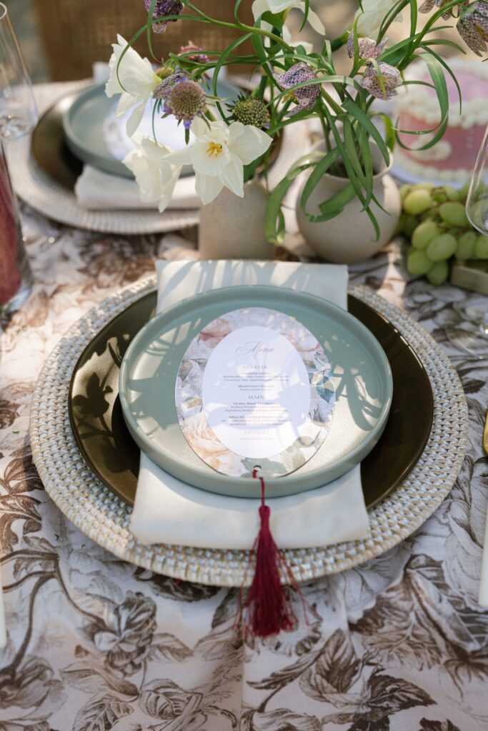An elegant outdoor tablescape featuring a floral‑patterned linen, wooden chairs with white cushions, layered place settings, burgundy‑tied napkins, lush garden‑inspired floral arrangements, and tiered cakes, styled as part of a refined San Diego wedding package celebration.