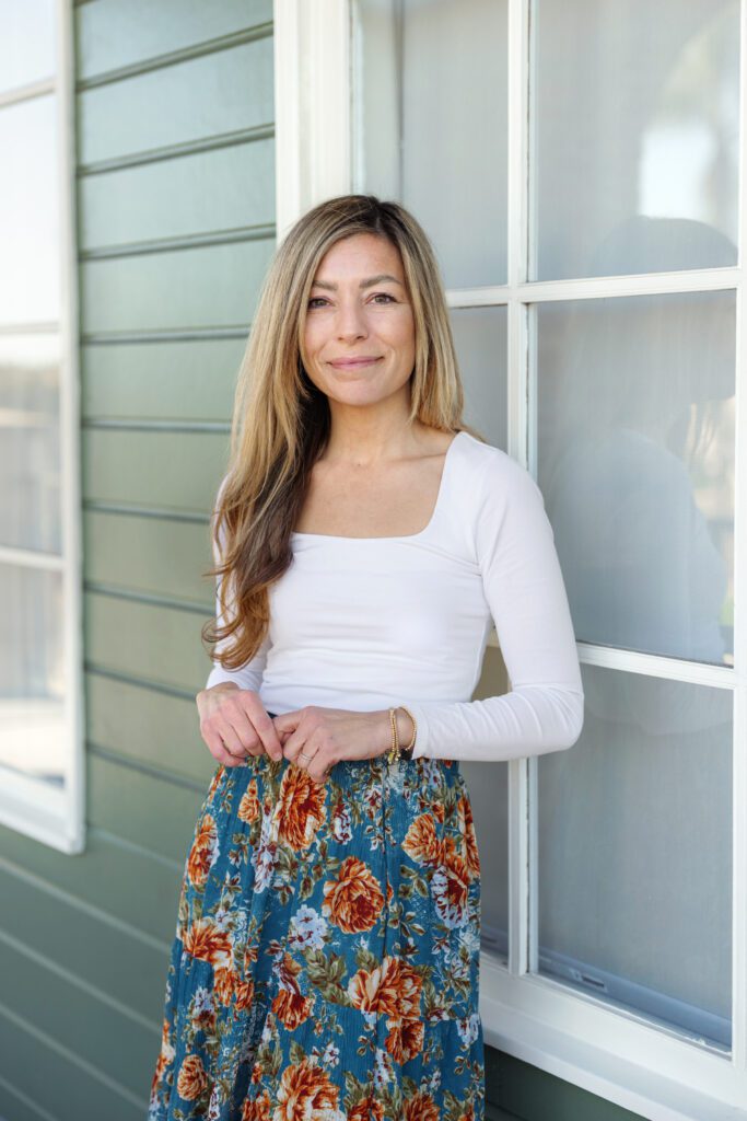 A wedding photographer standing outside a green‑sided building in a white long‑sleeve top and floral skirt, preparing to capture wedding day moments. 