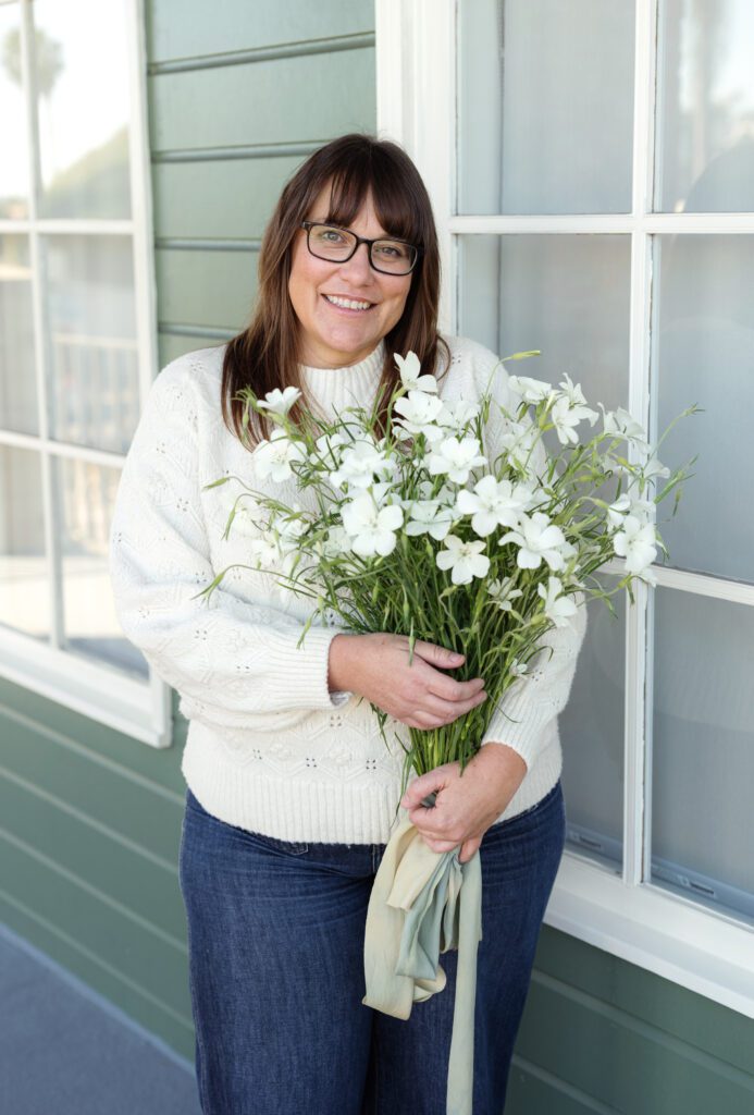 A florist standing outside a green‑sided building, holding a bouquet of white flowers tied with soft ribbons, capturing the organic, garden‑inspired style. 