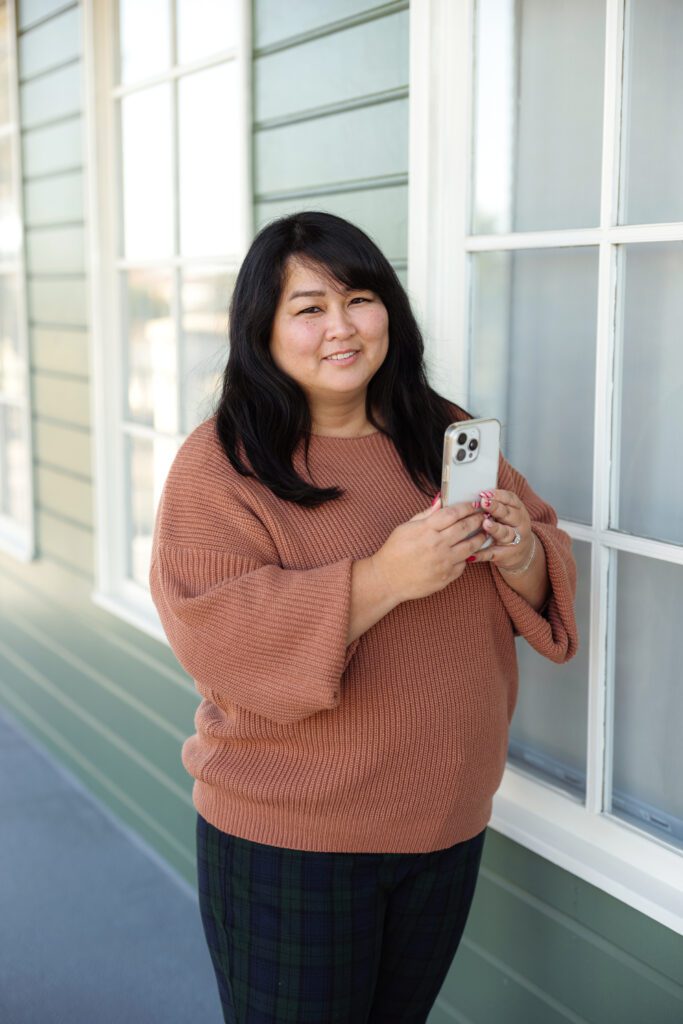 A content creator standing outside a green‑sided building in a rust sweater and plaid pants, capturing social content on her phone. 