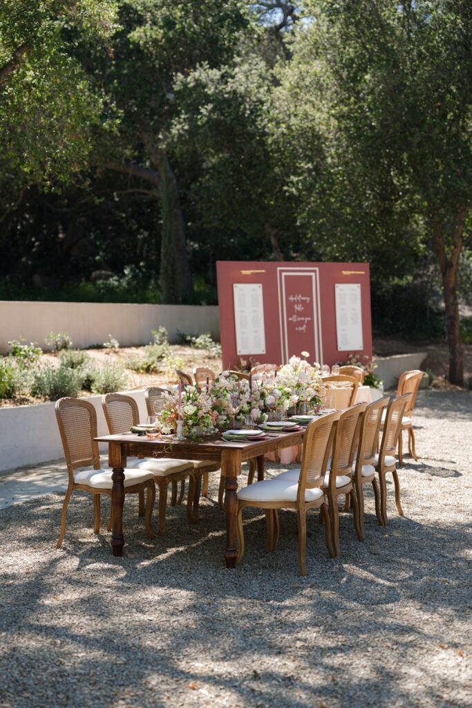 A long wooden reception table styled with pink and white floral centerpieces, elegant place settings, and mauve seating‑chart displays in the background, designed as part of an elevated San Diego wedding package experience.