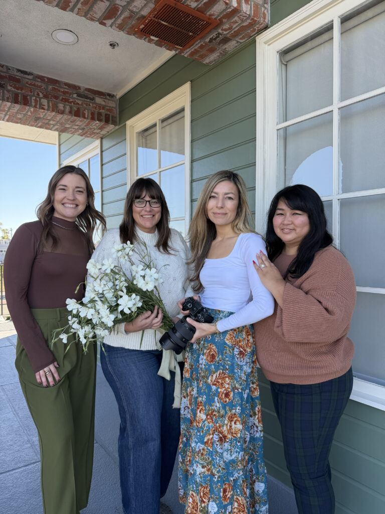 Four creative wedding professionals standing together on a porch, including a planner, photographer, florist, and content creator, holding flowers and a camera as they collaborate for a cohesive San Diego wedding package experience.