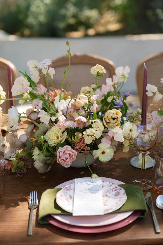  
A garden‑inspired tablescape featuring pastel florals, pink‑tinted glassware, floral‑patterned plates, and a printed menu, styled as part of an elevated San Diego wedding package